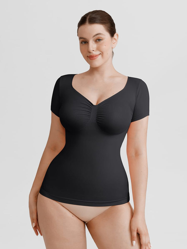 A woman modeling a black short-sleeve bodysuit with a ruched sweetheart neckline, paired with beige underwear, against a solid light gray background.