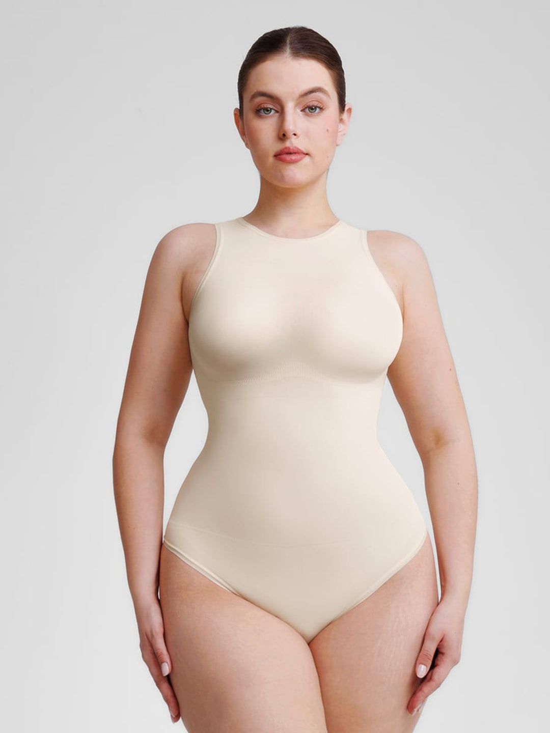 A woman modeling a seamless, high-neck, sleeveless shapewear bodysuit in a light beige shade against a solid light gray background.