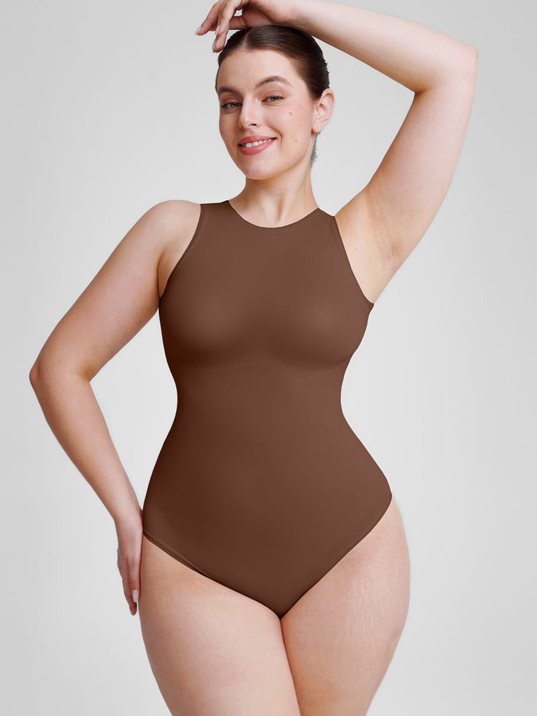 A woman modeling a sleeveless, high-neck, high-cut brown bodysuit against a light gray studio background.