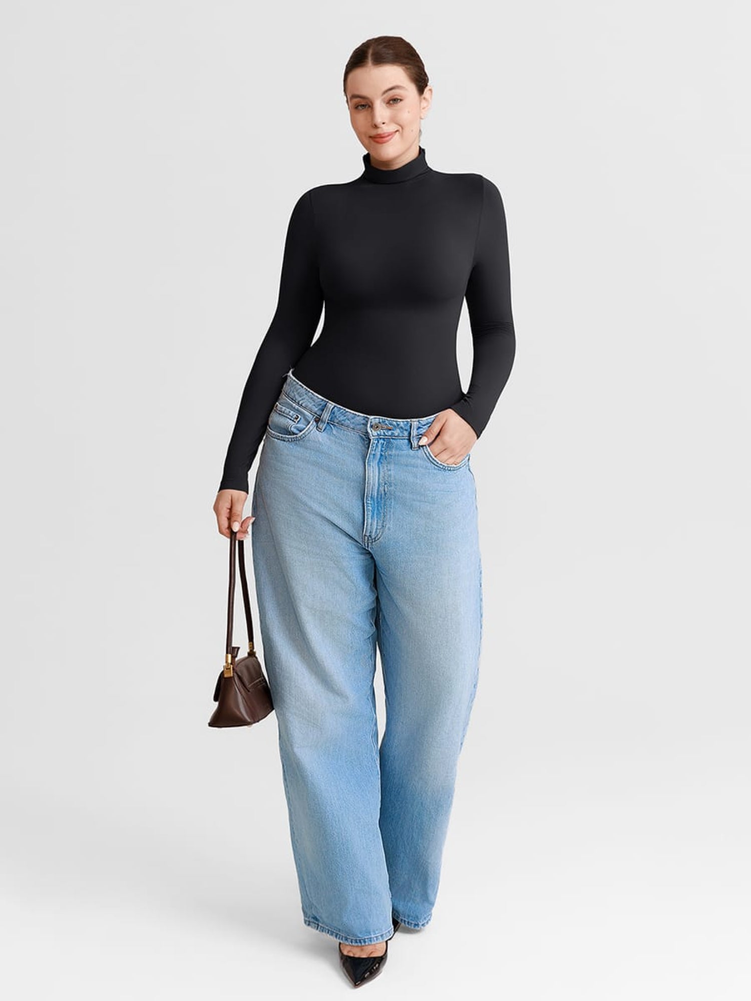 A woman modeling a black long-sleeve mock neck top with light-wash wide-leg jeans, holding a small brown handbag against a light gray studio background.