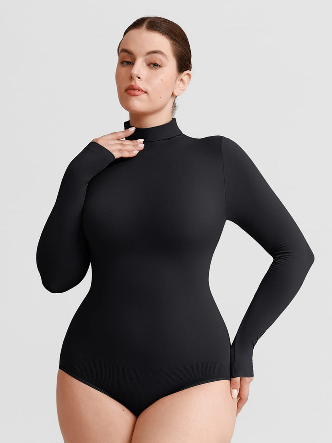A woman modeling a black long-sleeve mock neck bodysuit against a solid light gray background.