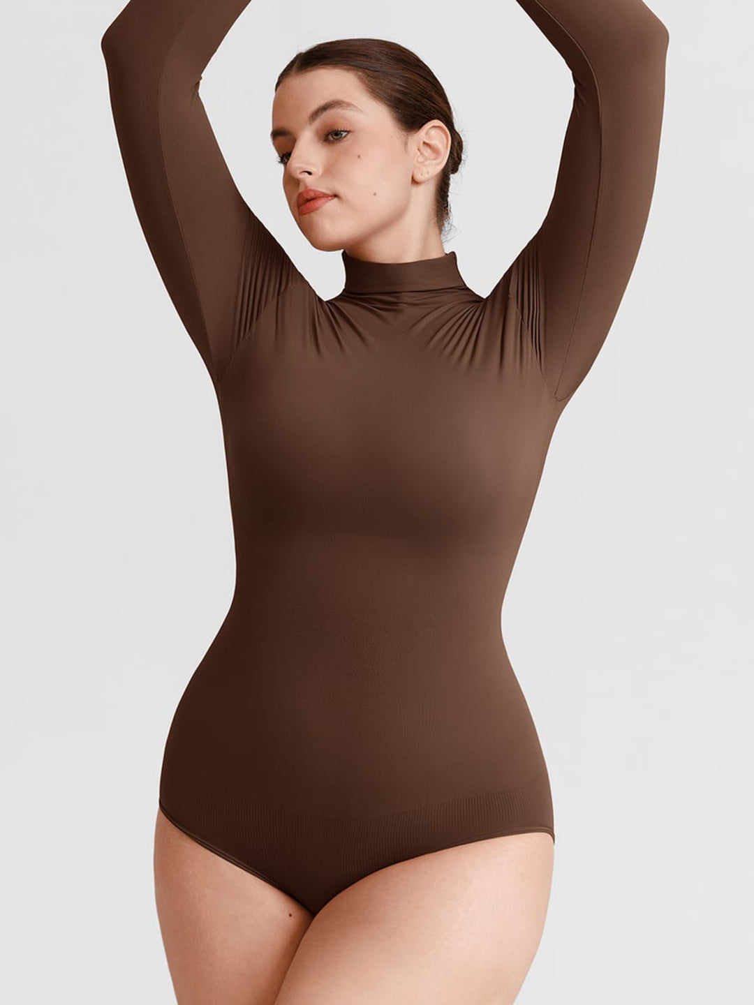 A woman modeling a long-sleeve, high-neck bodysuit in a solid dark brown color, posed with her arms raised against a light gray studio background.