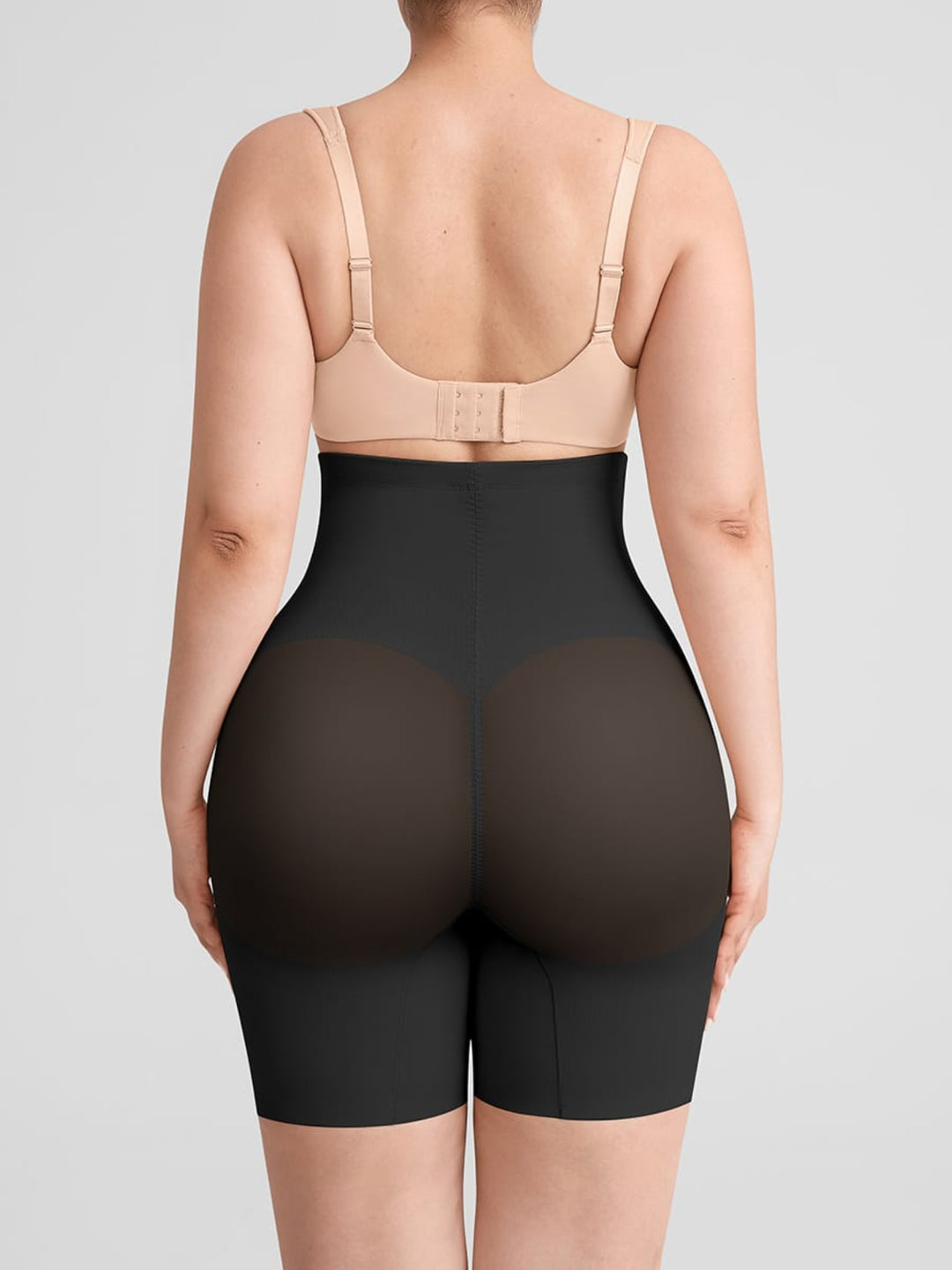 Rear view of a model wearing high-waisted black shapewear briefs and a beige bra against a light gray studio background.