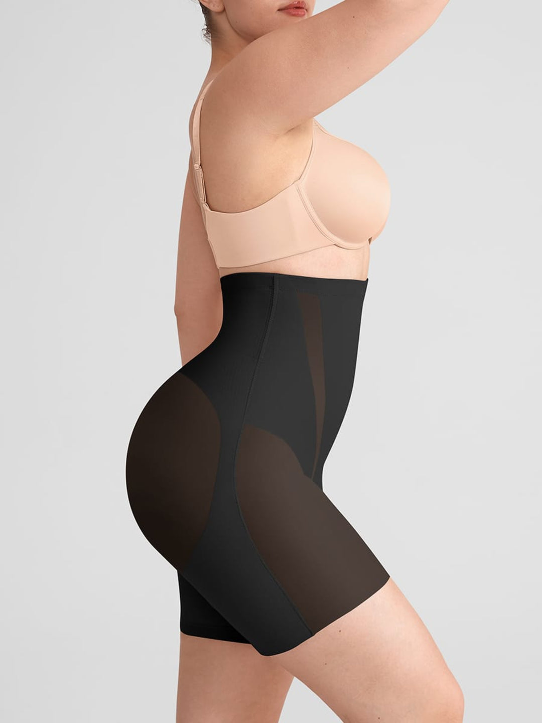 A model shown from the side wearing high-waisted black shapewear shorts with sheer mesh side panels over a nude/beige bra, against a light gray studio background.