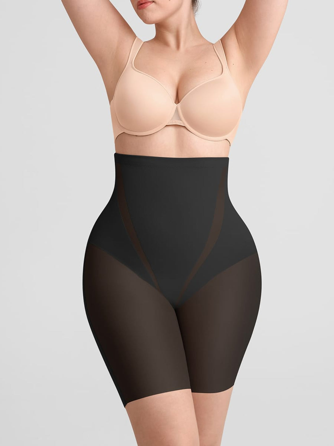 A model wearing high-waisted black shapewear briefs with sheer side panels, paired with a beige bra, against a solid light gray background.