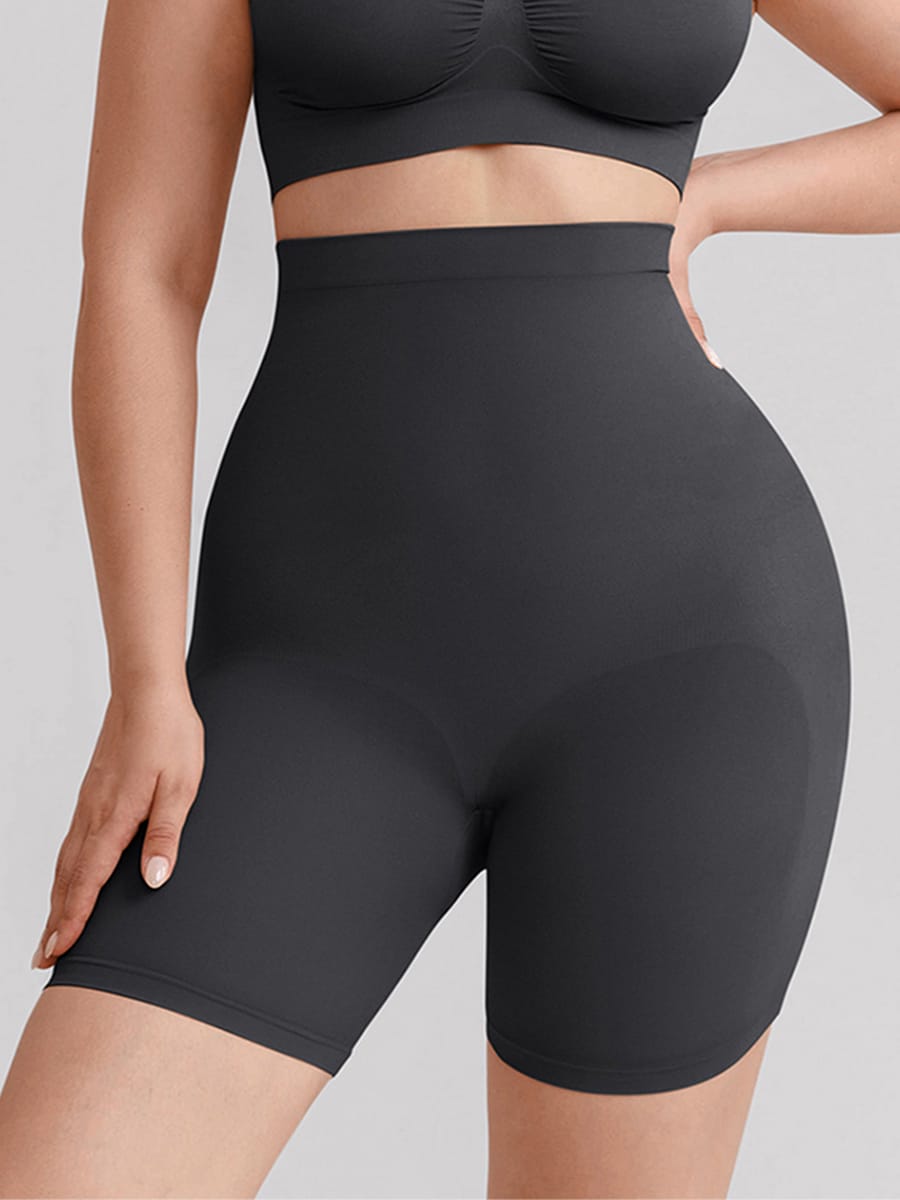 A close-up of a model wearing high-waisted black shapewear shorts and a matching black sports bra against a light gray studio background.