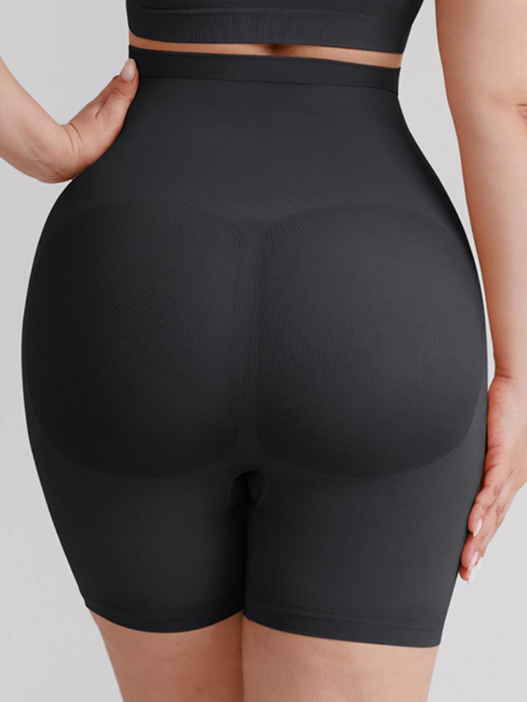 Rear view of a person wearing black high-waisted shapewear shorts against a light gray studio background.
