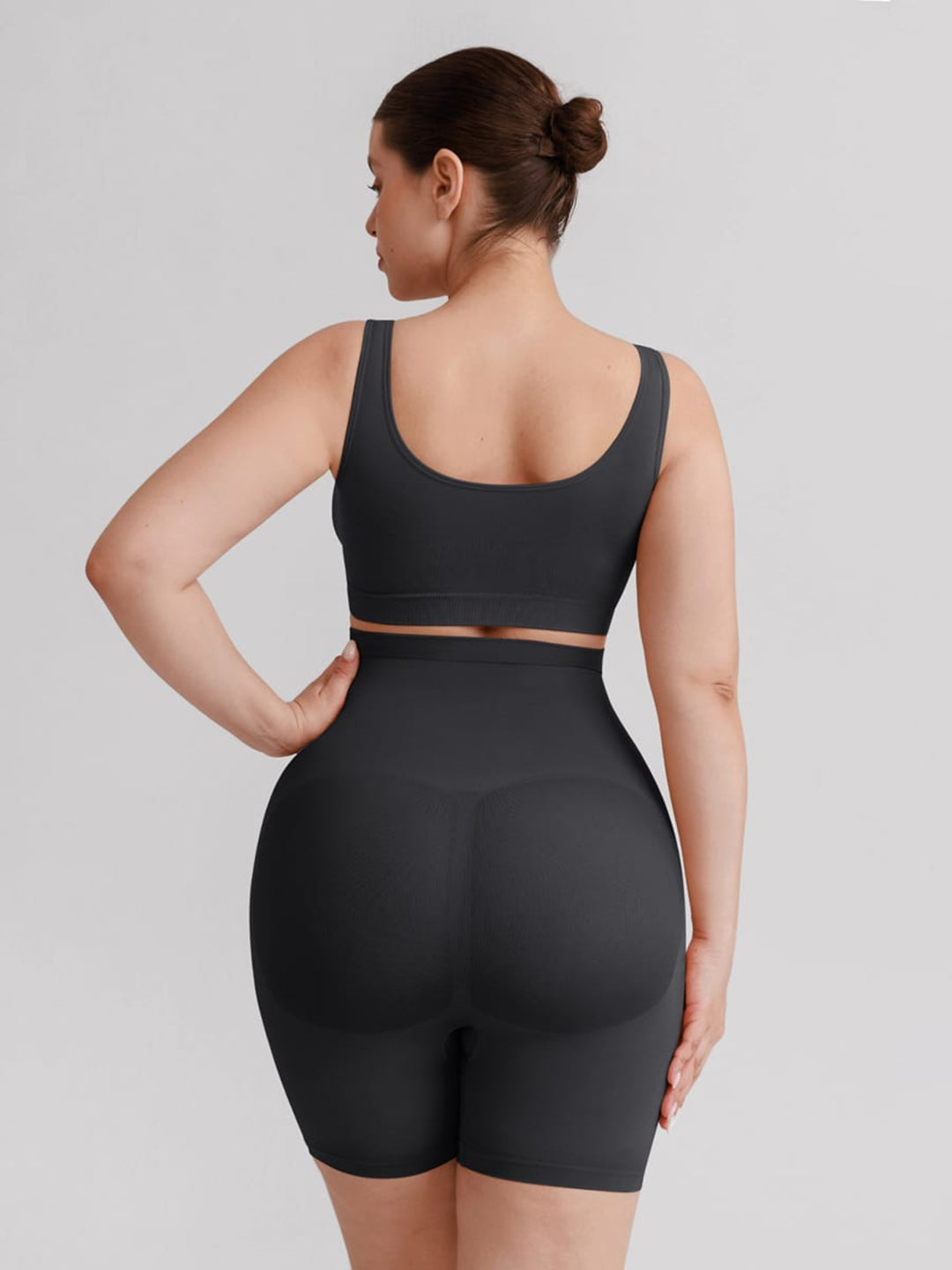 A woman shown from the back wearing a matching dark gray athletic set consisting of a sports bra and high-waisted biker shorts, against a solid light gray background.
