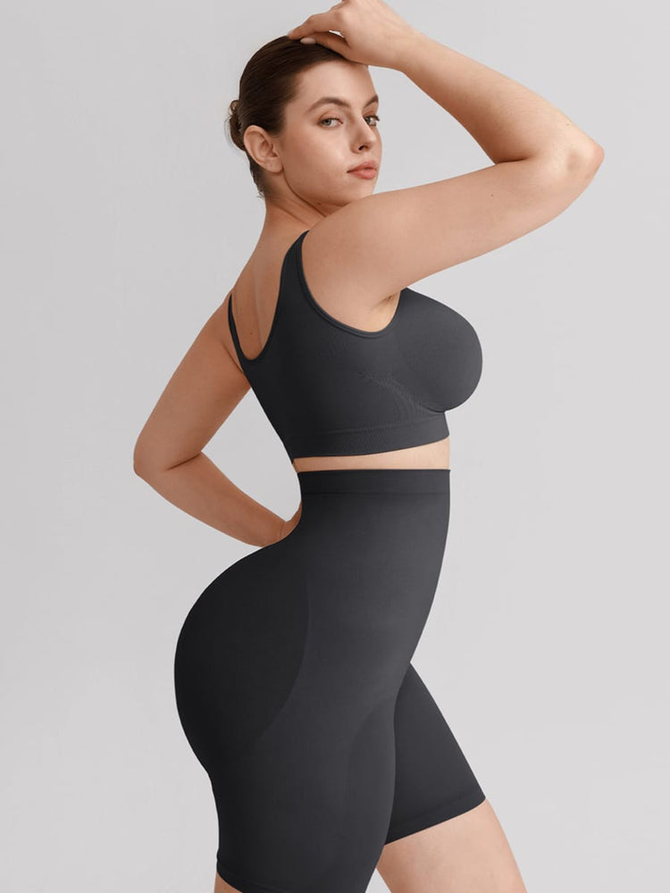 A woman modeling a matching black athletic set consisting of a sports bra and high-waisted shapewear shorts, photographed against a solid light gray studio background.