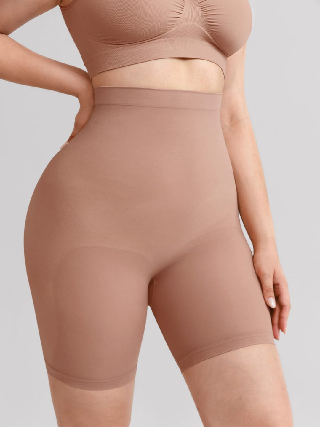 A close-up of a model wearing high-waisted shapewear shorts in a solid nude-beige color, paired with a matching bralette, against a solid light gray background.