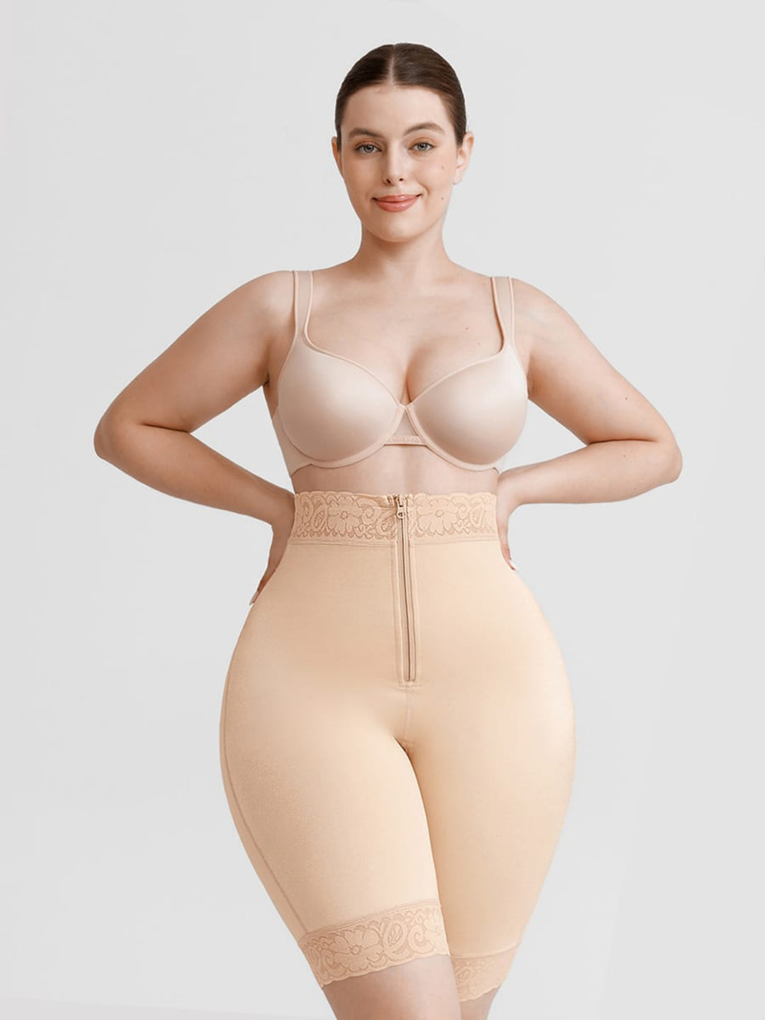 A woman modeling a beige shapewear brief with a lace waistband and zipper, paired with a matching bra, against a light gray studio background.