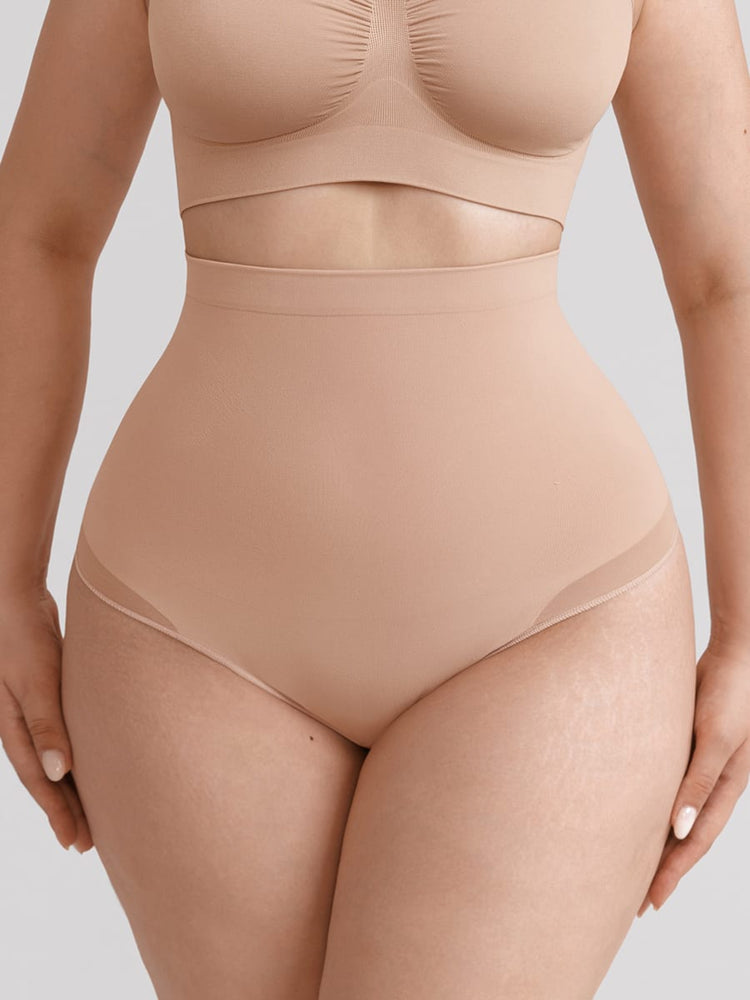 A close-up of a model wearing a high-waisted shapewear brief in a nude beige color, paired with a matching bralette, against a solid light gray background.