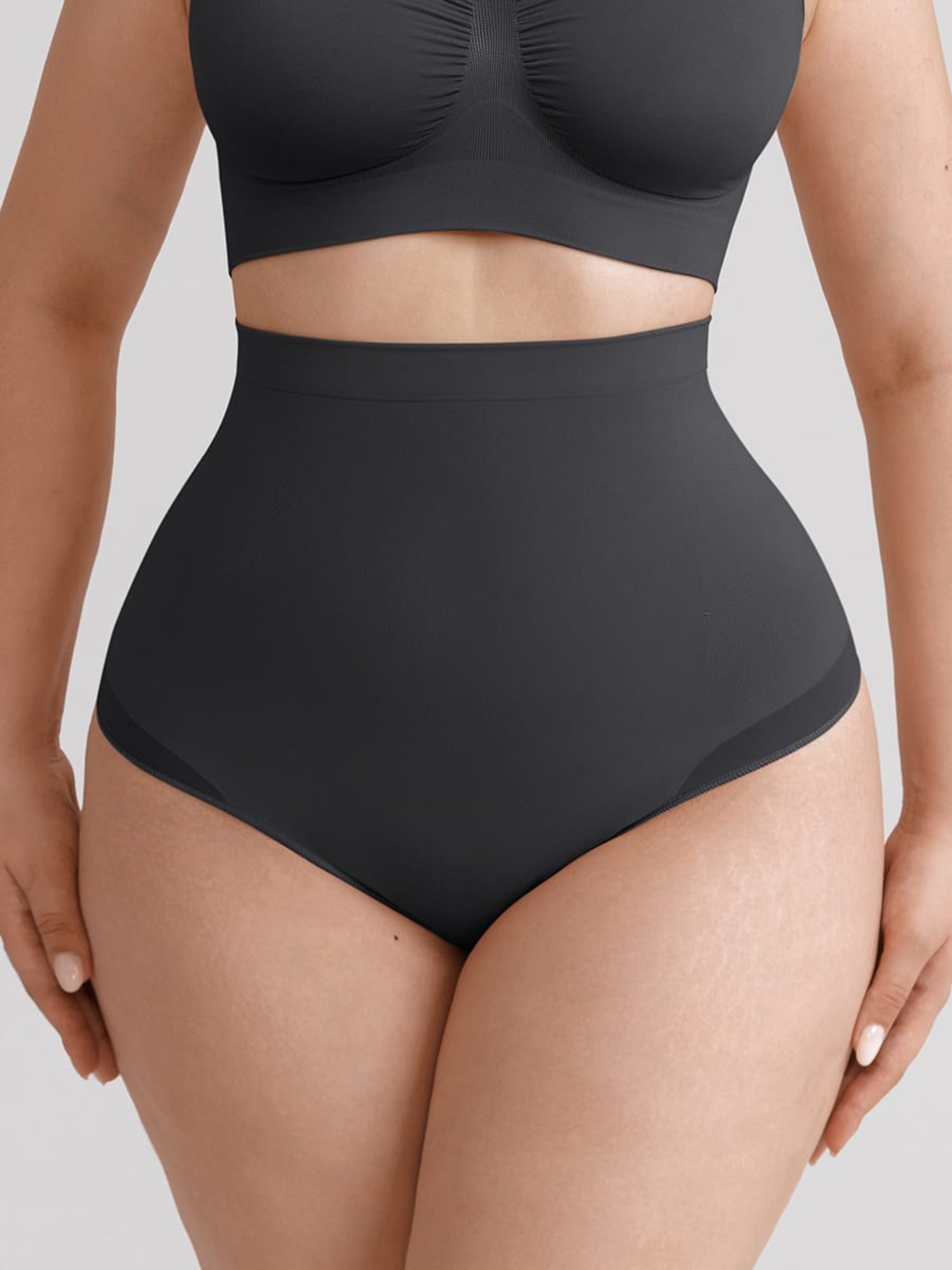 A close-up of a model wearing a dark charcoal gray two-piece swimsuit, featuring a high-waisted brief style bottom and a matching cropped top, set against a solid light gray background.