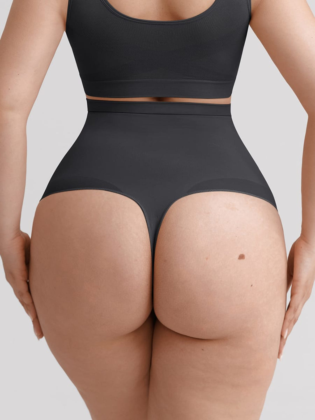 Rear view of a model wearing a black sports bra and high-waisted black briefs against a light gray background.