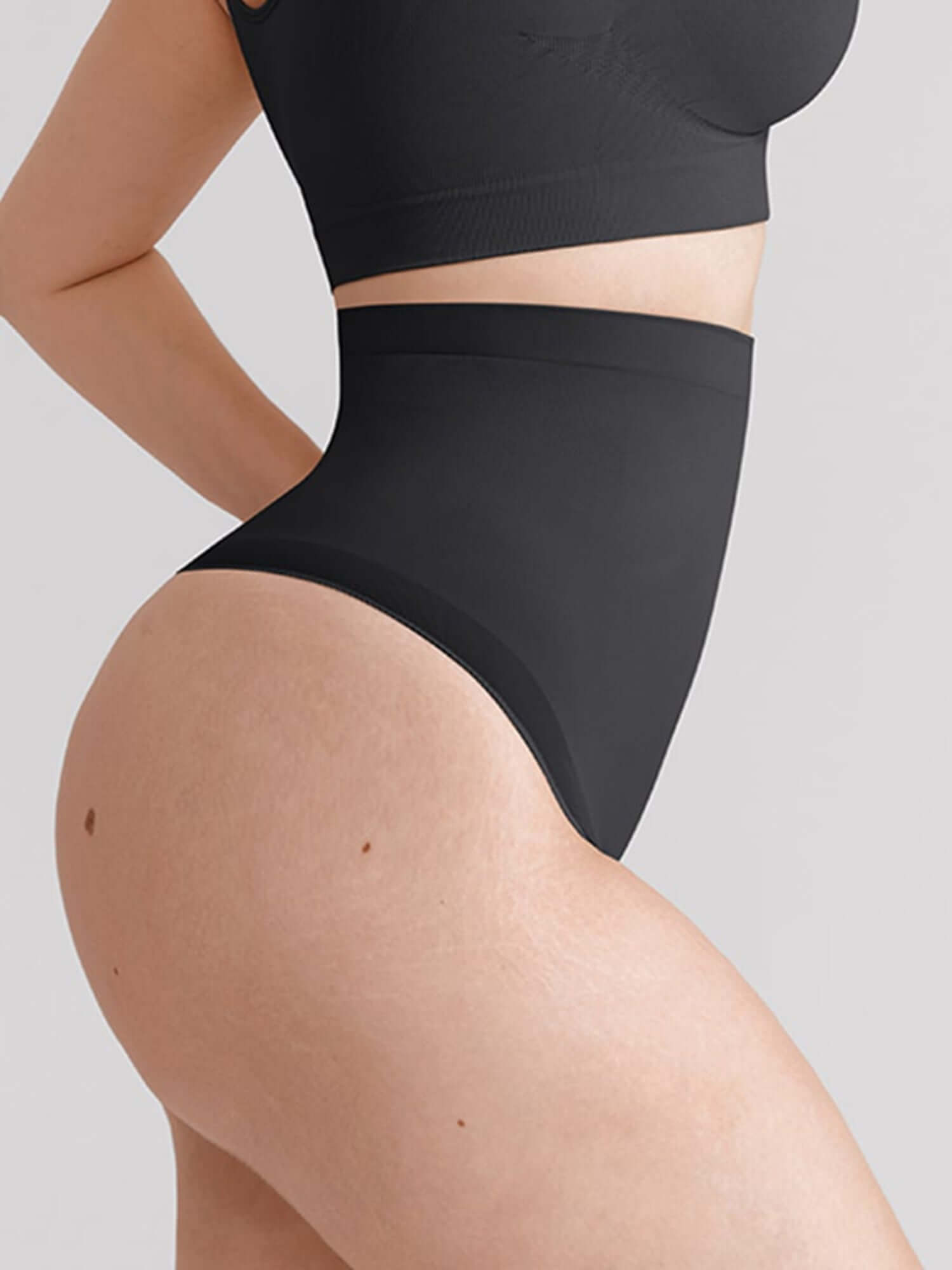 Close-up of a model wearing black high-waisted briefs and a matching black sports bra against a light gray background.