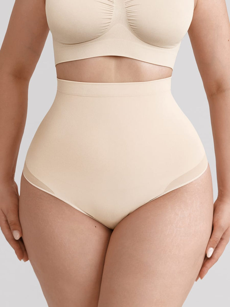 A close-up of a model wearing a two-piece shapewear set in a light beige/cream color, featuring a bralette top with a ruched center and high-waisted briefs, set against a solid gray background.