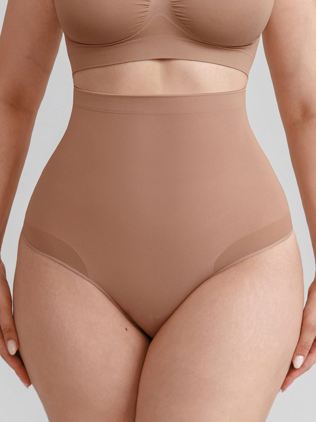 A close-up of a model wearing high-waisted shapewear briefs in a warm nude beige color, paired with matching bra top.