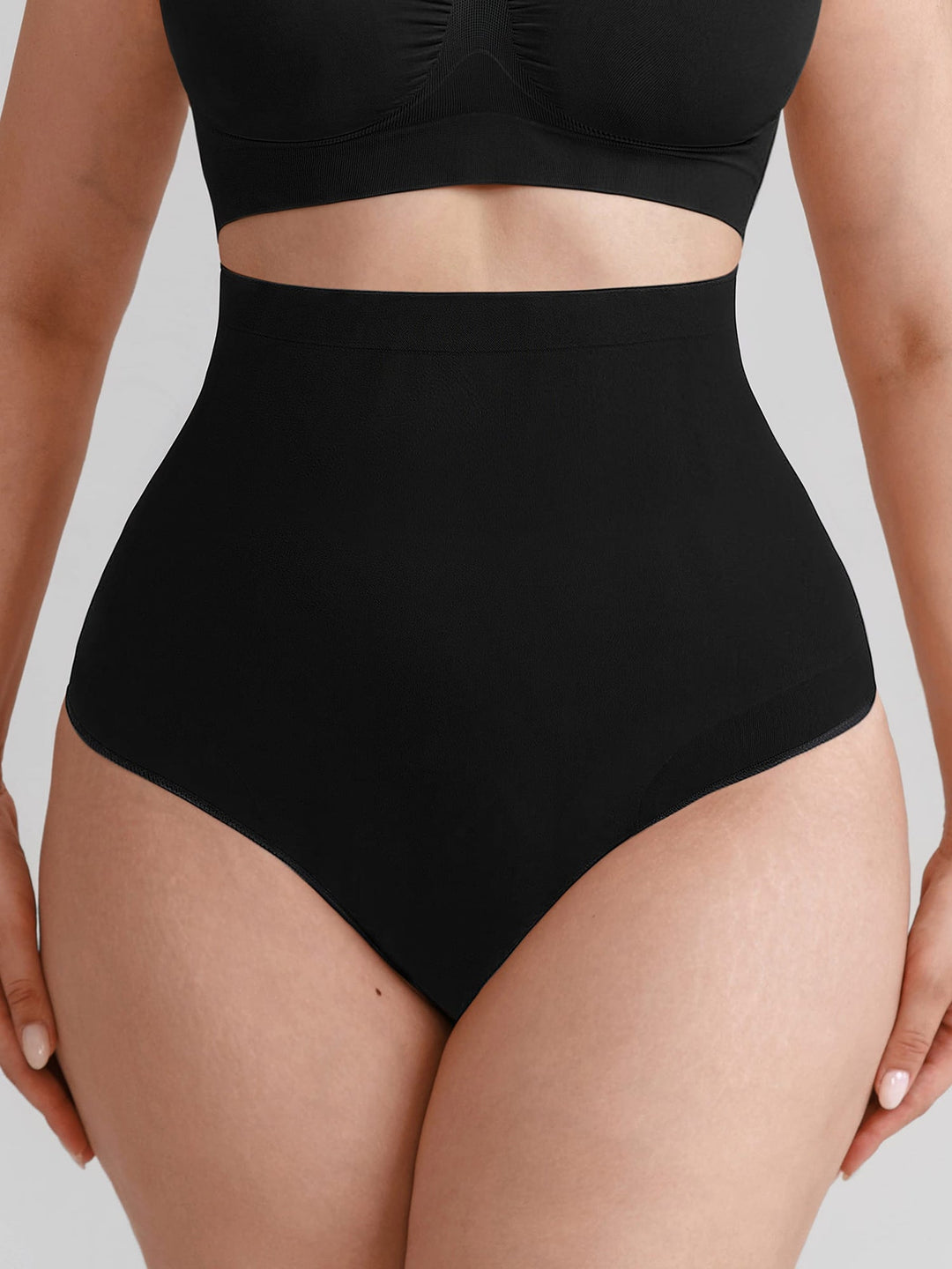 A close-up of a model wearing a solid black high-waisted brief and a matching black crop top against a light gray studio background.