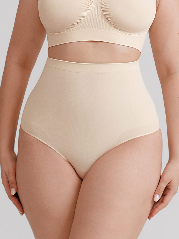 A close-up of a model wearing high-waisted seamless briefs and a matching bralette in a light beige nude color, set against a solid light gray background.