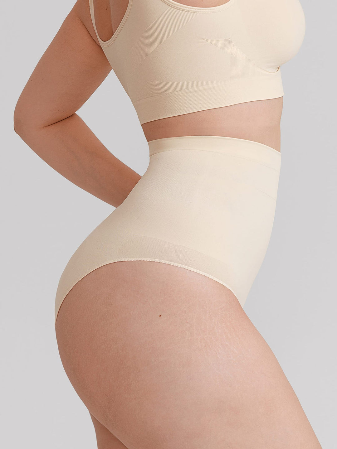 A close-up of a model wearing a matching beige two-piece lingerie set, featuring a high-waisted brief and a cropped tank top, against a solid light gray background.