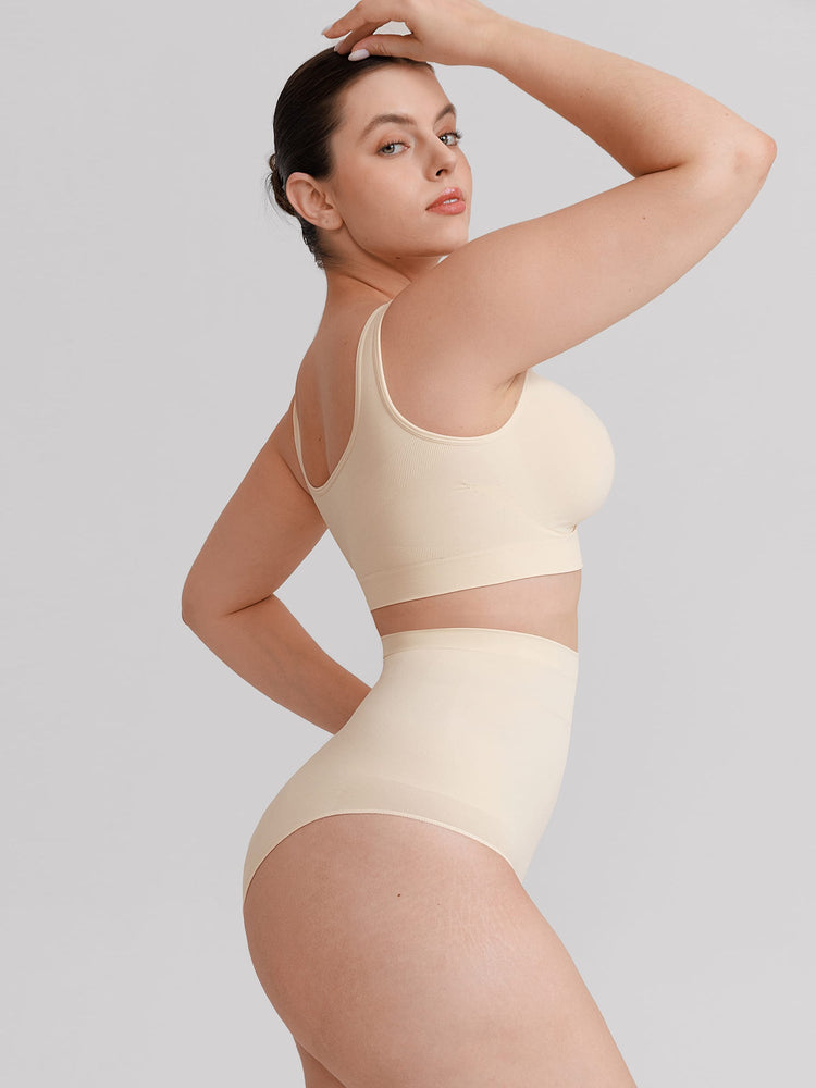 A woman modeling a matching beige two-piece lingerie set consisting of a cropped bralette and high-waisted briefs, posed against a solid light gray background.